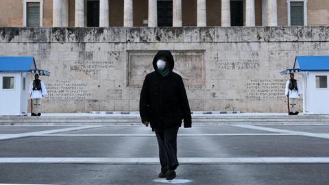 Eine Frau in schwarzer Kleidung trägt eine medizinische Maske und läuft damit über den Syntagma Platz in Athen. Im Hintergrund ist das Grab des Unbekannten Soldaten zu sehen.