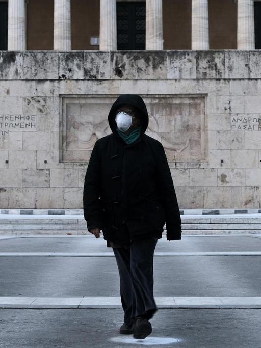 Eine Frau in schwarzer Kleidung trägt eine medizinische Maske und läuft damit über den Syntagma Platz in Athen. Im Hintergrund ist das Grab des Unbekannten Soldaten zu sehen.