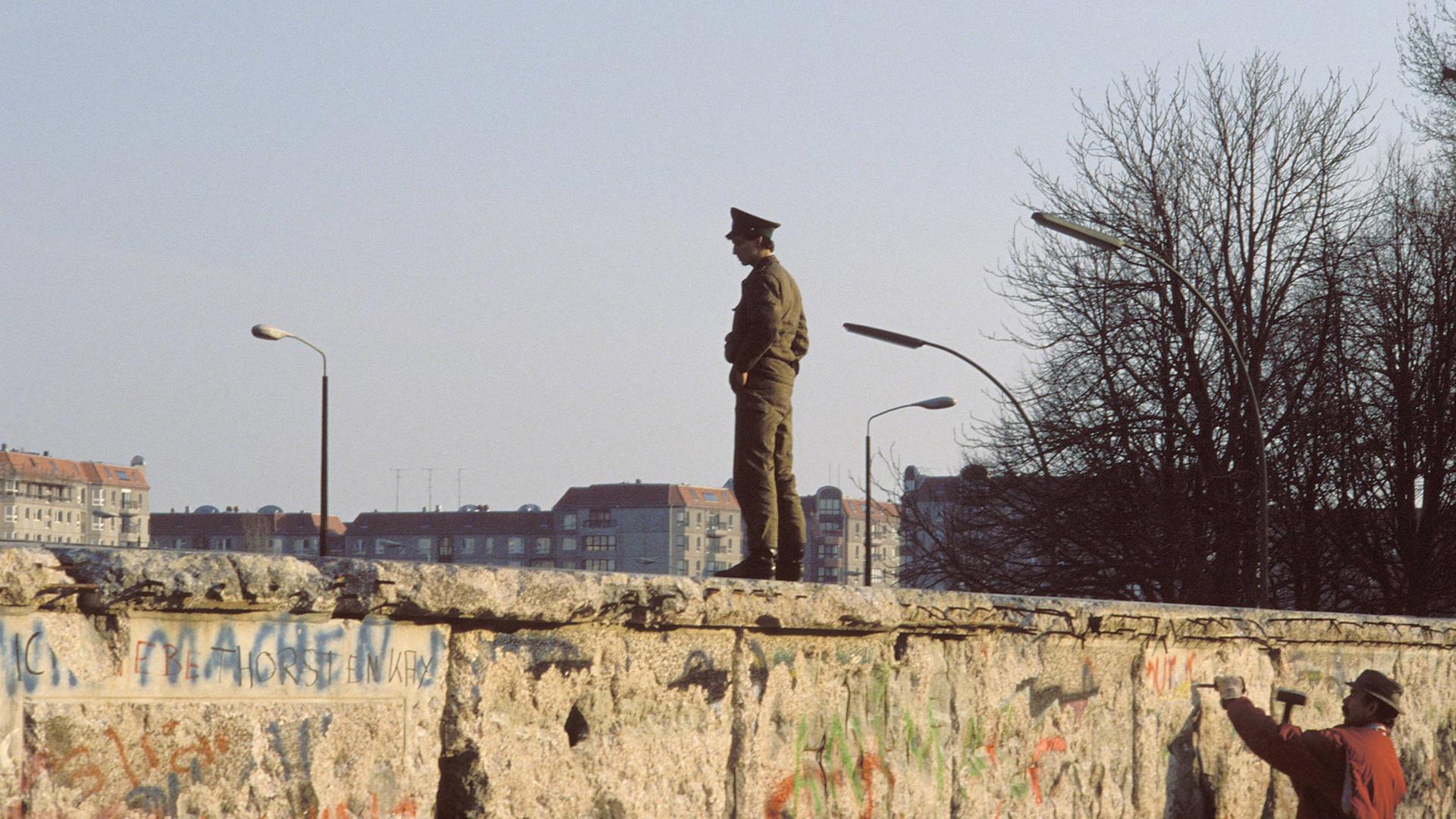 Ein Mann mit Hammer und Meisel im Wendejahr 19989/90 an der Berliner Mauer. Oben auf der Mauer steht ein DDR-Grenzposten.