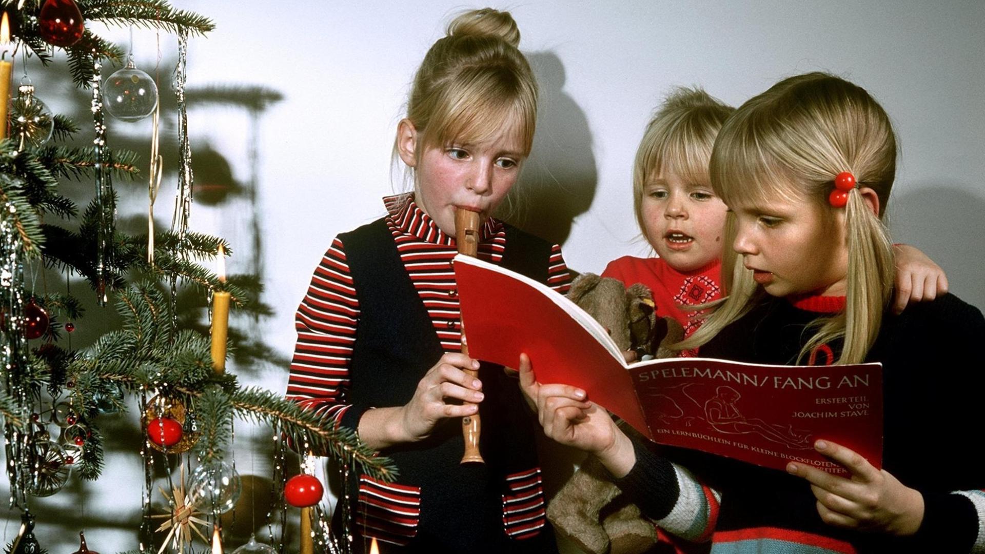 Zwei Mädchen stehen am Heiligen Abend neben dem Christbaum und singen Weihnachtslieder. Sie werden von ihrer Schwester auf der Blockflöte begleitet.