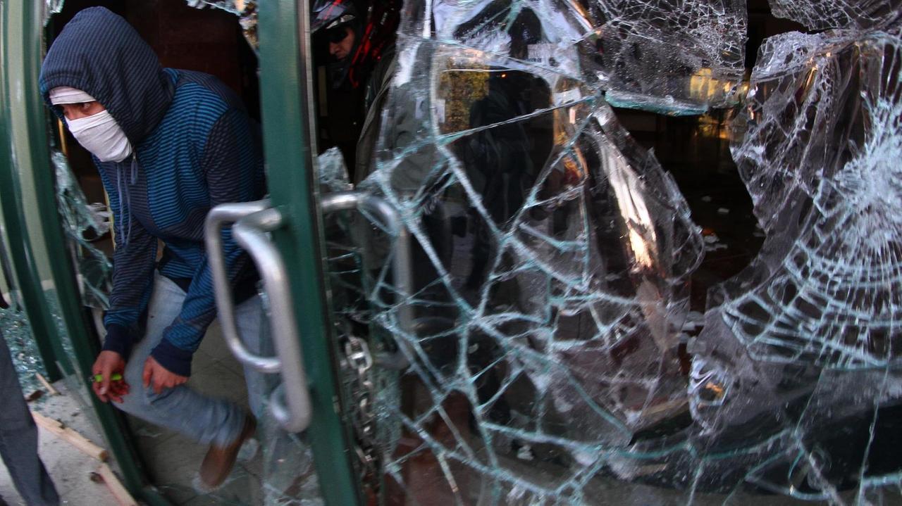 Ein Vermummter geht durch eine demolierte Glastür.