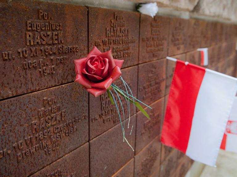 Gedenkstätte für die Opfer des Massenmords von Katyn. Eine polnische Flagge und eine rote Rose stecken an einer Mauer mit eingravierten Namen.