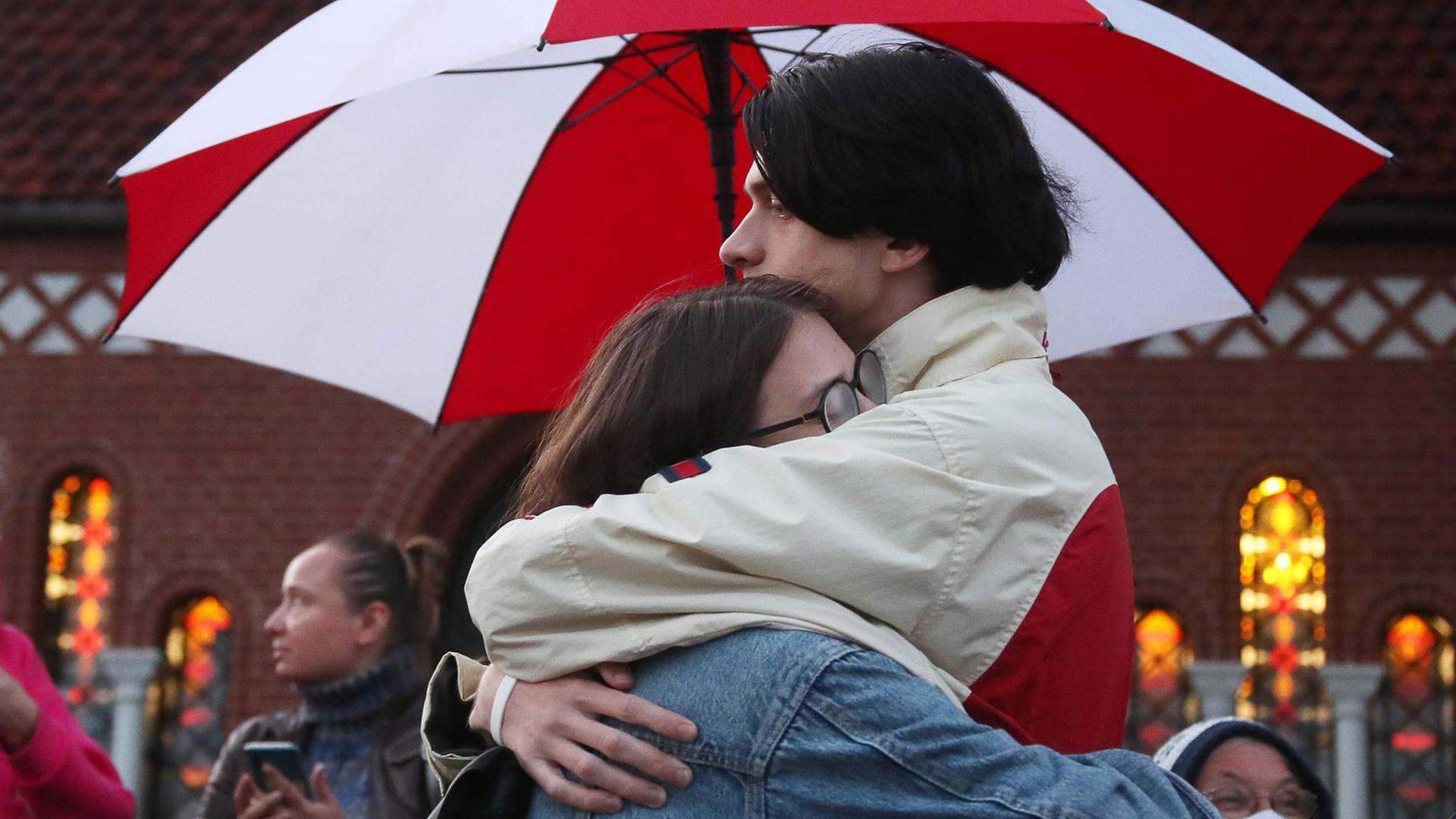 Zwei Demonstranten umarmen sich unter einem rot-weißen Regenschirm.