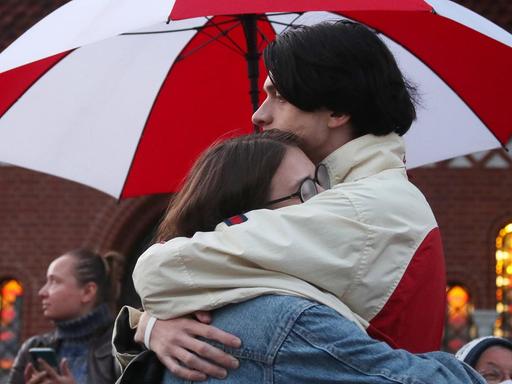 Zwei Demonstranten umarmen sich unter einem rot-weißen Regenschirm.