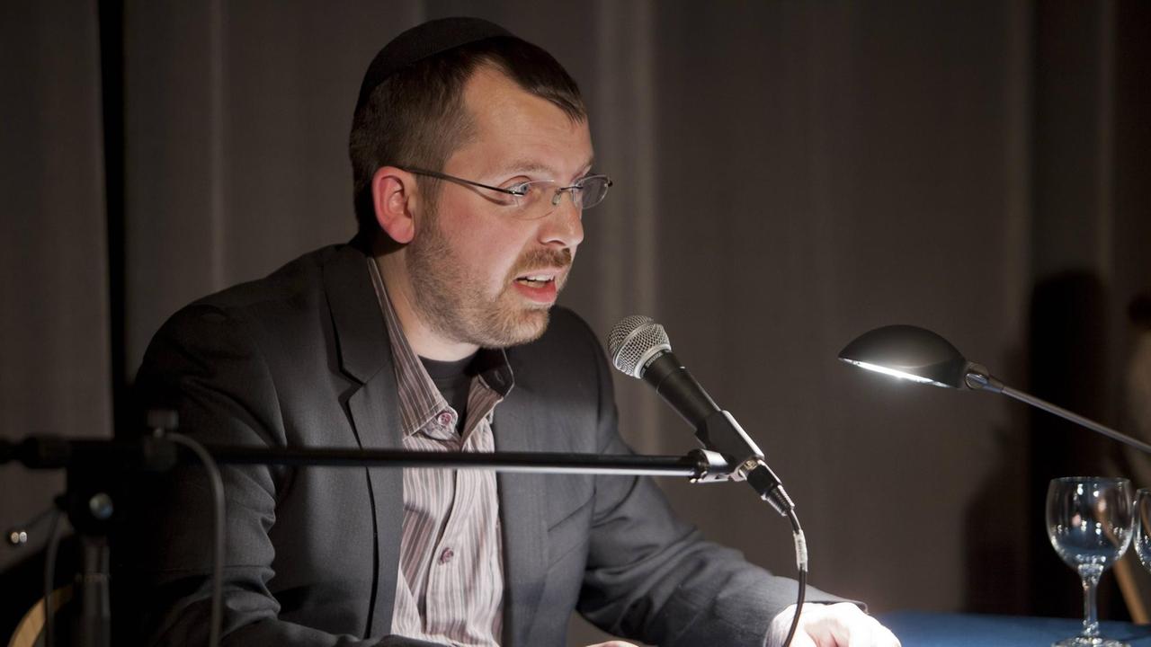 Der Autor Eliyah Havemann sitzt an einem Tisch mit einer kleinen Leselampe vor einem Mikrofon und hält auf dem Tisch einige Blätter Paper in den Händen, die er auf dem Tisch abgelegt hat. Der Autor Eliyah Havemann sitzt an einem Tisch mit einer kleinen Leselampe vor einem Mikrofon und hält auf dem Tisch einige Blätter Paper in den Händen, die er auf dem Tisch abgelegt hat.