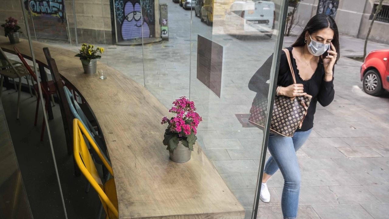 Eine junge Frau mit Coronamaske läuft in Barcelona an einem leeren Café vorbei. Eine junge Frau mit Coronamaske läuft in Barcelona an einem leeren Café vorbei.