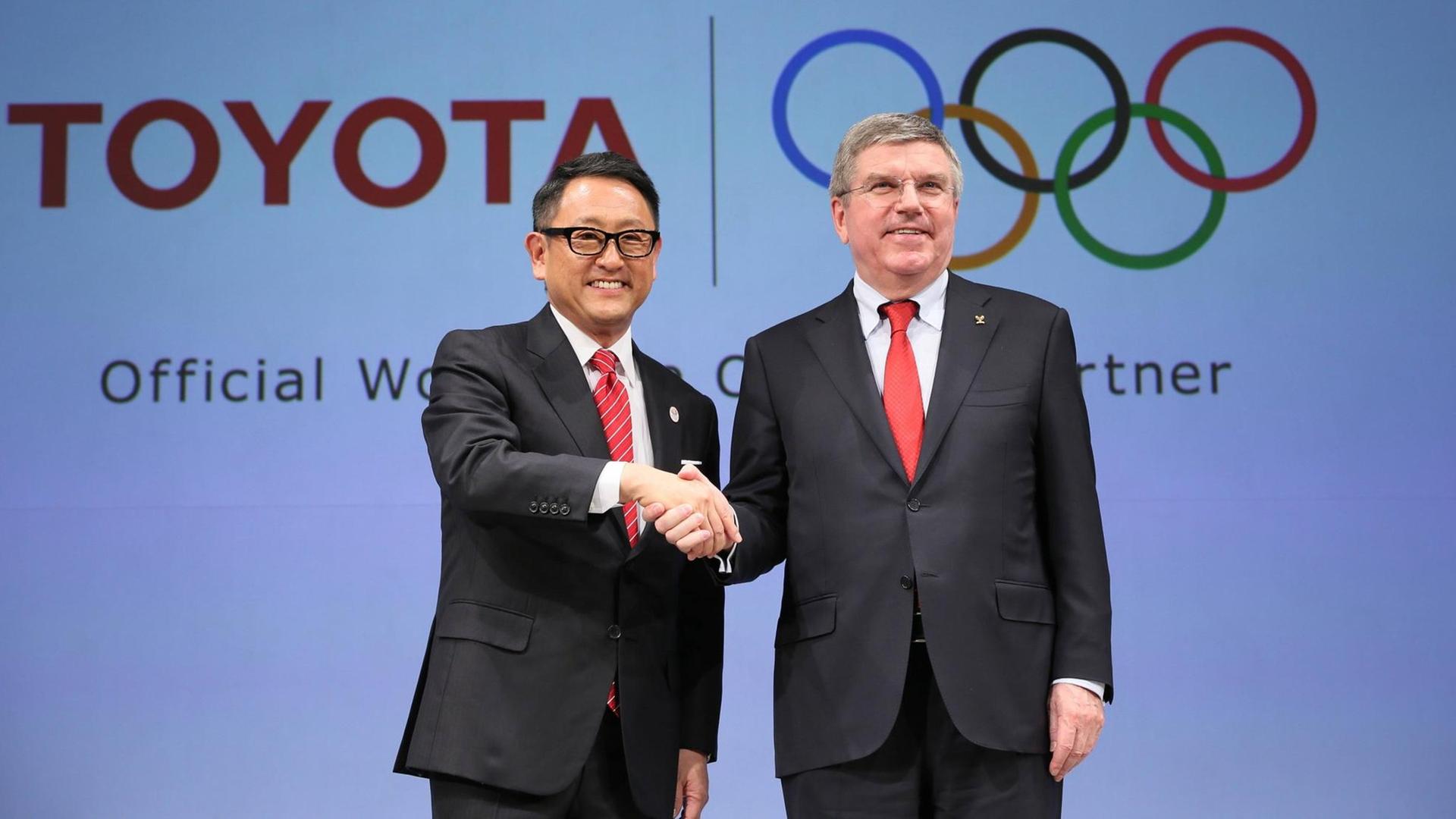 Toyota-Konzernchef Akio Toyoda und IOC-Präsident Thomas Bach schütteln sich die Hände, im Hintergrund die Logos von Toyota und den Olympischen Spielen Toyota-Konzernchef Akio Toyoda und IOC-Präsident Thomas Bach schütteln sich die Hände, im Hintergrund die Logos von Toyota und den Olympischen Spielen