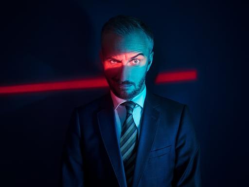 Jan Böhmermann blickt kritisch-spöttisch in die Kamera, eine Augenbraue hochgezogen. Er trägt Anzug und Krawatte und steht im Dunklen. Ein Spalt rotes Licht leuchtet auf seine Augenpartie und setzt diese so in den Fokus des Fotos.