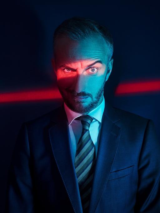 Jan Böhmermann blickt kritisch-spöttisch in die Kamera, eine Augenbraue hochgezogen. Er trägt Anzug und Krawatte und steht im Dunklen. Ein Spalt rotes Licht leuchtet auf seine Augenpartie und setzt diese so in den Fokus des Fotos.