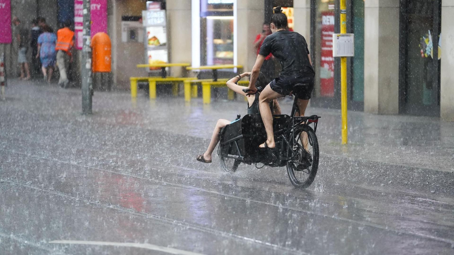 Junge Leute fahren mit einem Lastenfahrrad, während starker Regen auf die Berliner Friedrichstraße prasselt. Junge Leute fahren mit einem Lastenfahrrad, während starker Regen auf die Berliner Friedrichstraße prasselt.