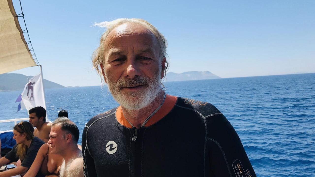Ein Mann mit weißem Haar und Bart steht mit Tauchanzug auf seinem Boot im Wasser. Ein Mann mit weißem Haar und Bart steht mit Tauchanzug auf seinem Boot im Wasser.