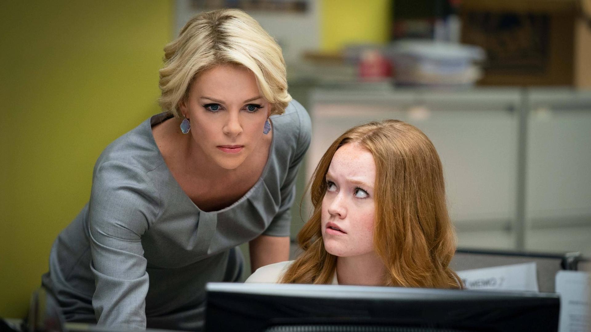 In ihrer Rolle als Megyn Kelly im Film "Bombshell" beugt sich die Schauspielerin Charlize Theron über einen Schreibtisch und schaut auf den Bildschirm einer Kollegin. In ihrer Rolle als Megyn Kelly im Film "Bombshell" beugt sich die Schauspielerin Charlize Theron über einen Schreibtisch und schaut auf den Bildschirm einer Kollegin.