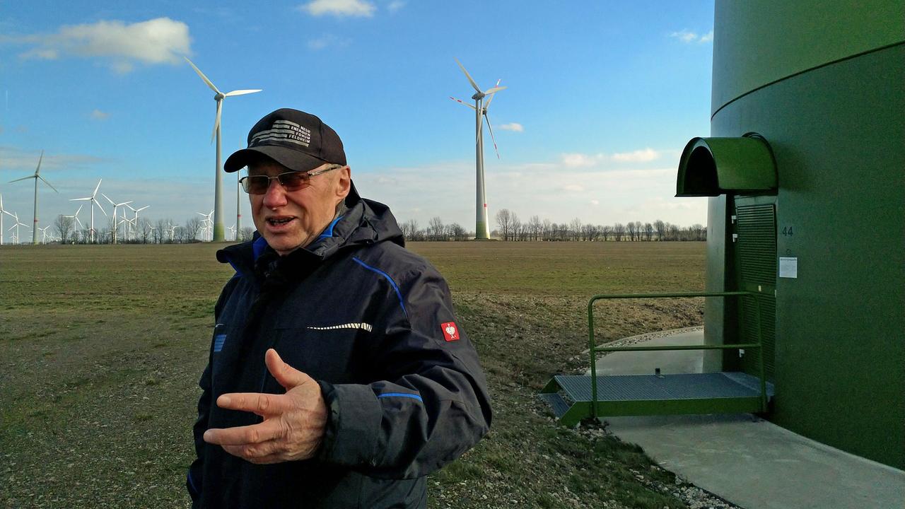Mit blauer Jacke und dunkler Baseballkappe steht Siegfried Kappert auf einer Wiese. Im Hintergrund Windräder. Mit blauer Jacke und dunkler Baseballkappe steht Siegfried Kappert auf einer Wiese. Im Hintergrund Windräder.