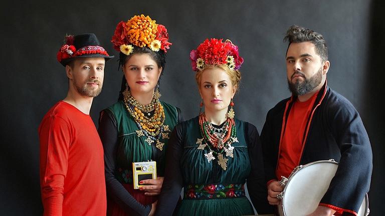 Zwei Männer und zwei Frauen in bunten Kleidern mit bunten Hüten aus Blumen stehen nebeneinander und blicken in die Kamera. Sie halten unterschiedliche Musikinstrumente in der Hand. Zwei Männer und zwei Frauen in bunten Kleidern mit bunten Hüten aus Blumen stehen nebeneinander und blicken in die Kamera. Sie halten unterschiedliche Musikinstrumente in der Hand.