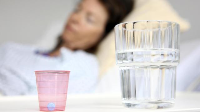 Eine Patientin liegt in einem Krankenbett, Medikament und Wasserglas auf dem Nachttisch im Bildvordergrund