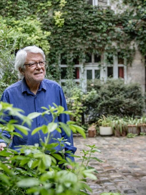 Georges Aperghis steht in einem begrünten Innenhof und lächelt — sein Blick geht an der Kamera vorbei.