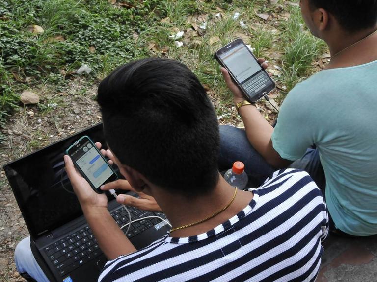 In einem Park in Havanna sitzen zwei junge Männer, die mit ihren Handys und einem Laptop im Internet sind.