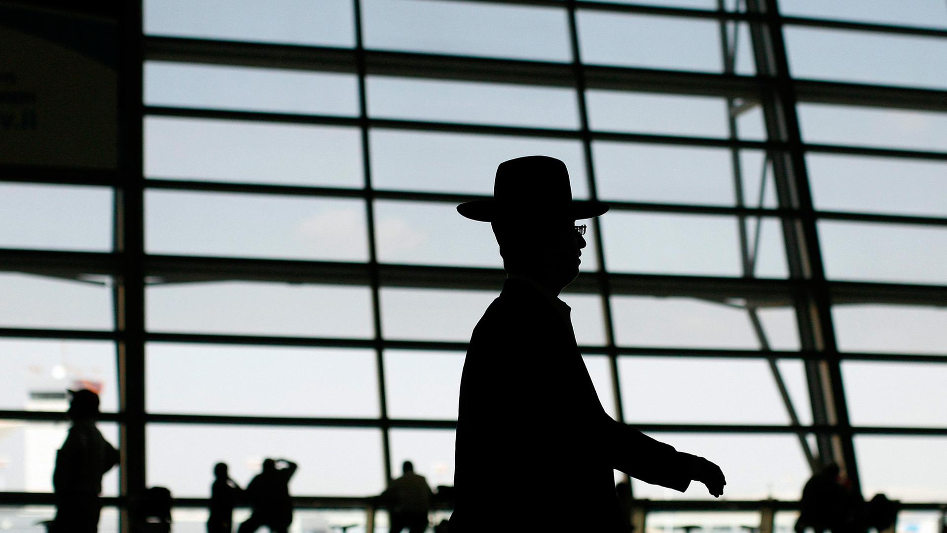 Abflugshalle des Flughafens Ben Gurion in Israel: durch die einfallende Sonne liegen die Streben der Glasfront und die vorübergehenden Menschen im Schatten, markant im Vordergrund ist die Silhouette eines orthodoxen Juden zu sehen Abflugshalle des Flughafens Ben Gurion in Israel: durch die einfallende Sonne liegen die Streben der Glasfront und die vorübergehenden Menschen im Schatten, markant im Vordergrund ist die Silhouette eines orthodoxen Juden zu sehen