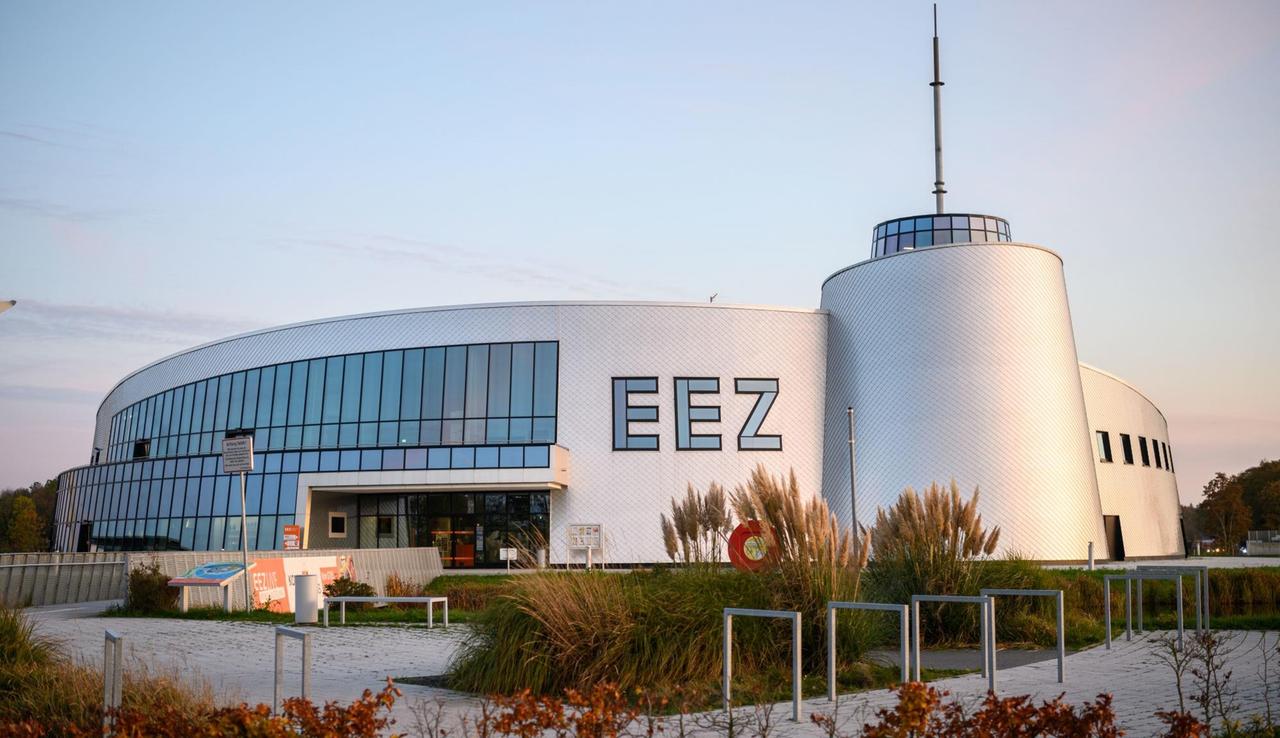 Blick auf das Gebäude des Enercon Besucherzentrums im EEZ (Energie-, Bildungs- und Erlebniszentrum). Das Gebäude ist rund und hat silbrig erscheinende Außenwände, EEZ steht in großen Buchstaben darauf. 