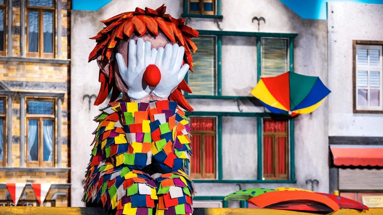 Eine Clowns-Figur sitzt mit den Händen vor dem Gesicht auf einem Rosenmontagswagen, links unten ist ein Modell eines Coronavirus zu sehen, im Hintergrund Modelle von Häusern Eine Clowns-Figur sitzt mit den Händen vor dem Gesicht auf einem Rosenmontagswagen, links unten ist ein Modell eines Coronavirus zu sehen, im Hintergrund Modelle von Häusern