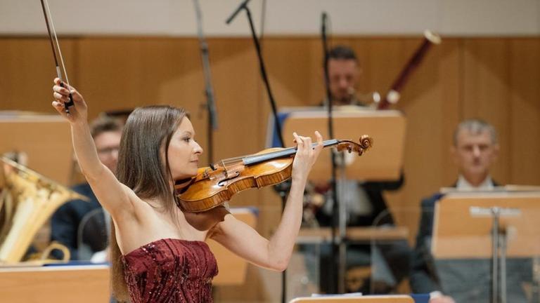 Eine Geigerin steht vor einem Orchester und schwingt ihren Bogen weit nach oben. Eine Geigerin steht vor einem Orchester und schwingt ihren Bogen weit nach oben.
