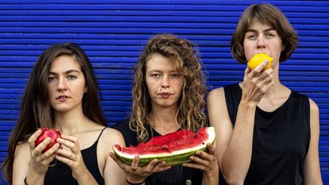 Drei Frauen stehen vor einem blauen Rolladen. Sie halten verschiedene Früchte in ihren Händen.