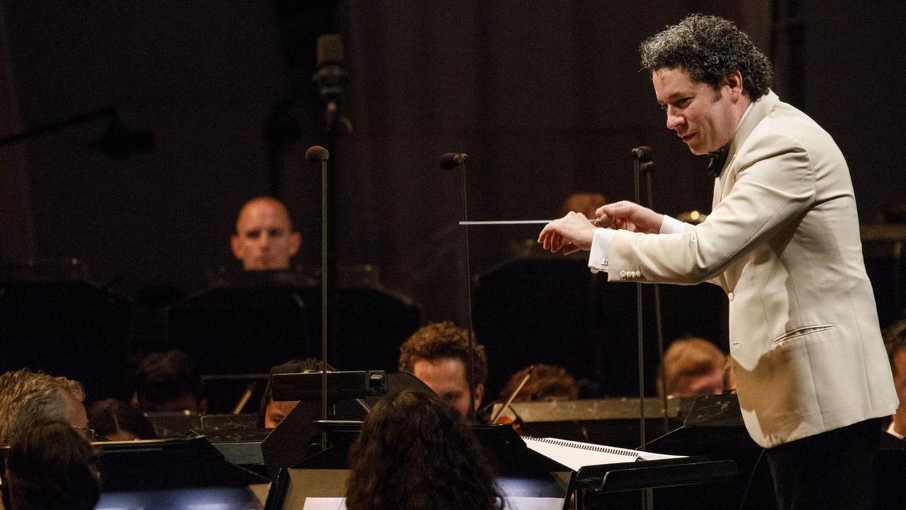 Gustavo Dudamel am Dirigentenpult. Er trägt eine helle Jacke und hebt sich damit vom dunklen, fast schwarzen Hintergrund ab. Gustavo Dudamel am Dirigentenpult. Er trägt eine helle Jacke und hebt sich damit vom dunklen, fast schwarzen Hintergrund ab.