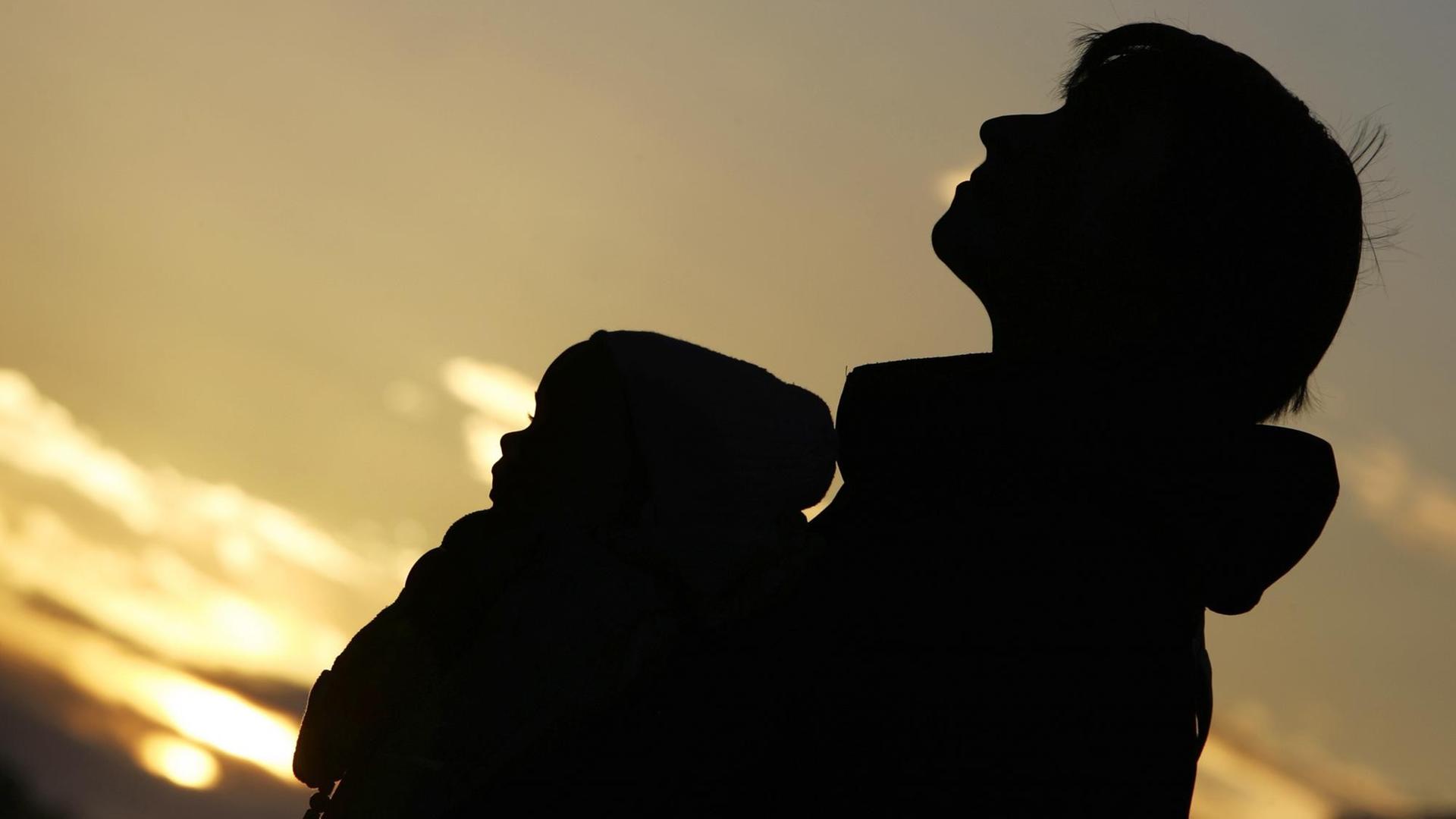 Ein Frau trägt bei Sonnenuntergang einen Säugling in einem Tragegurt vor der Brust Ein Frau trägt bei Sonnenuntergang einen Säugling in einem Tragegurt vor der Brust