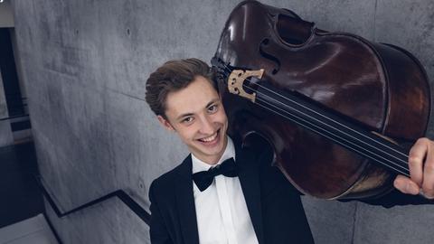 Der Cellist Sebastian Fritsch mit geschultertem Cello vor grauer Betonwand.