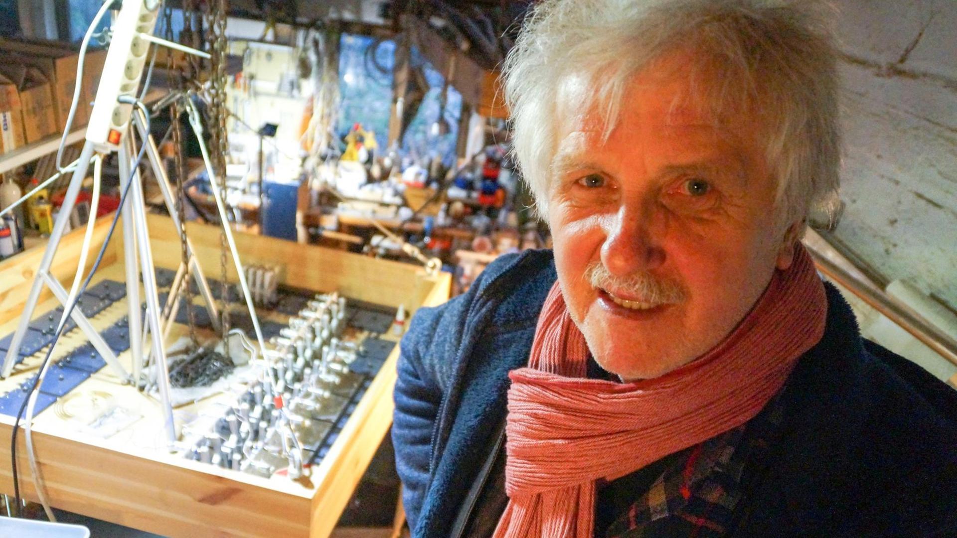 Ein Mann mit weißen Haaren und weißem Schnäuzer steht im Vordergrund des Bildes. Hinter ihm stehen lauter ungewöhnliche Geräte, Musikinstrumente, die er selbst gebaut hat. Ein Mann mit weißen Haaren und weißem Schnäuzer steht im Vordergrund des Bildes. Hinter ihm stehen lauter ungewöhnliche Geräte, Musikinstrumente, die er selbst gebaut hat.