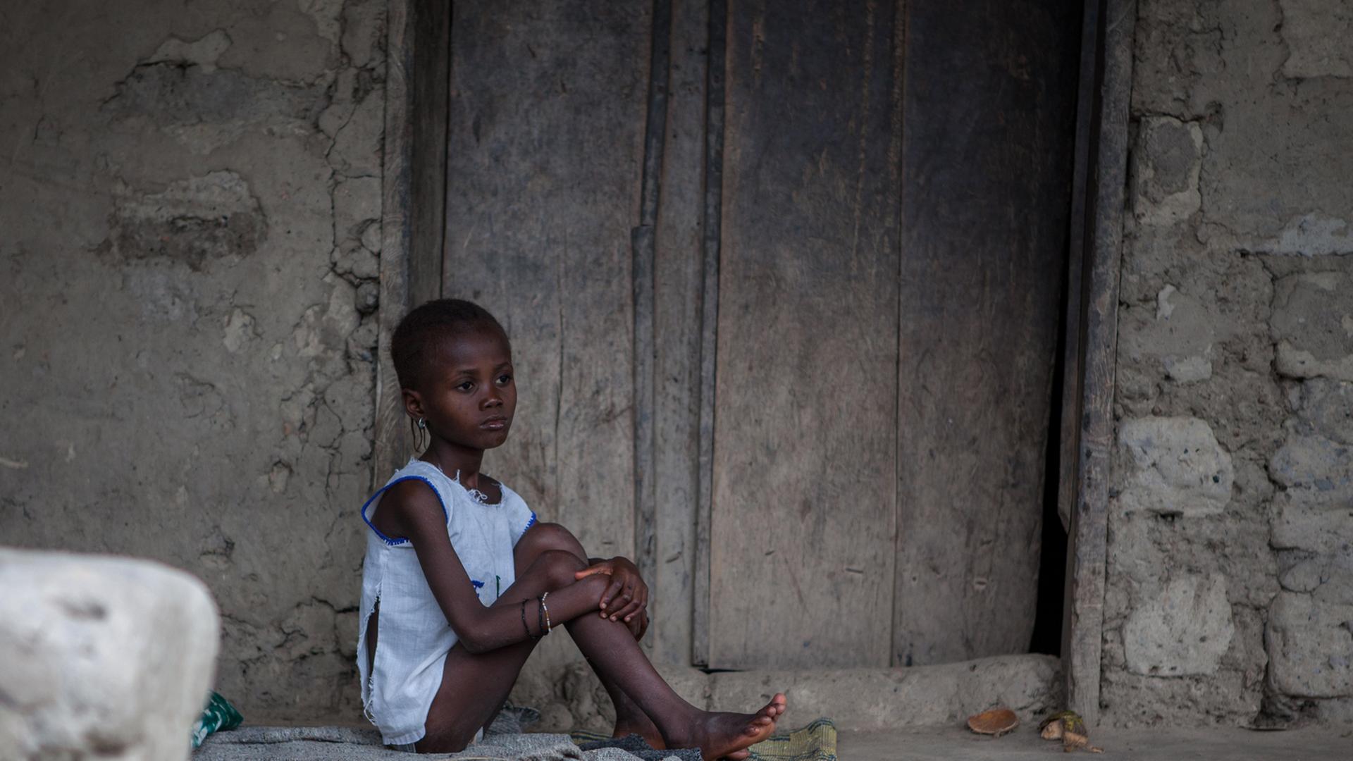 Ein kleines Mädchen sitzt vor einem Haus auf dem Boden. Sie könnte mit Ebola infiziert sein.