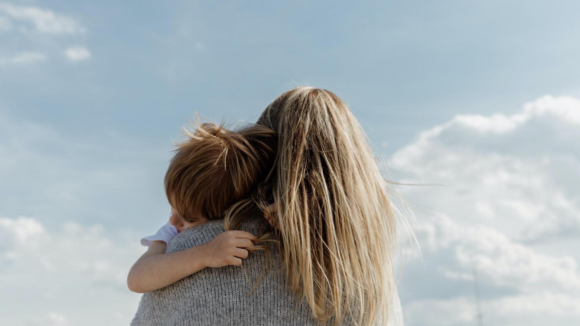 Eine Person mit langen blonden Haaren trägt ein Kind auf dem Arm. Sie sind von schräg unten aufgenommen, im Hintergrund erstreckt sich der blaue Himmel. Eine Person mit langen blonden Haaren trägt ein Kind auf dem Arm. Sie sind von schräg unten aufgenommen, im Hintergrund erstreckt sich der blaue Himmel.