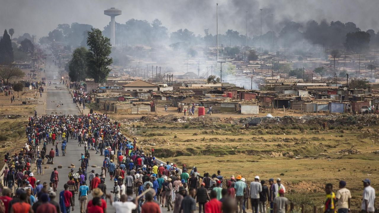 Bewaffnete Menschen rennen auf eine Siedlung zu, aus der Rauch aufsteigt.