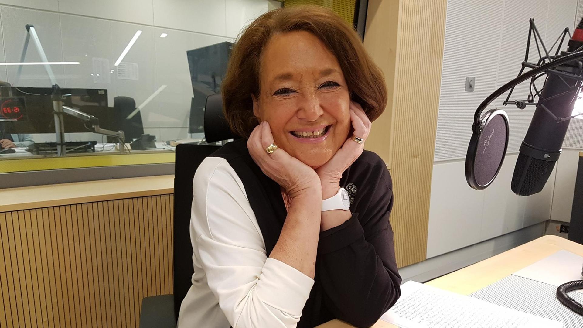 Eine ältere Dame sitzt in einem Radiostudio und lächelt in die Kamera, während sie den Kopf auf die Hände gestützt hat. Eine ältere Dame sitzt in einem Radiostudio und lächelt in die Kamera, während sie den Kopf auf die Hände gestützt hat.