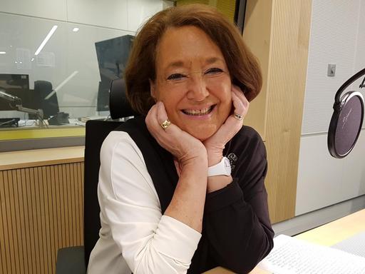 Eine ältere Dame sitzt in einem Radiostudio und lächelt in die Kamera, während sie den Kopf auf die Hände gestützt hat.