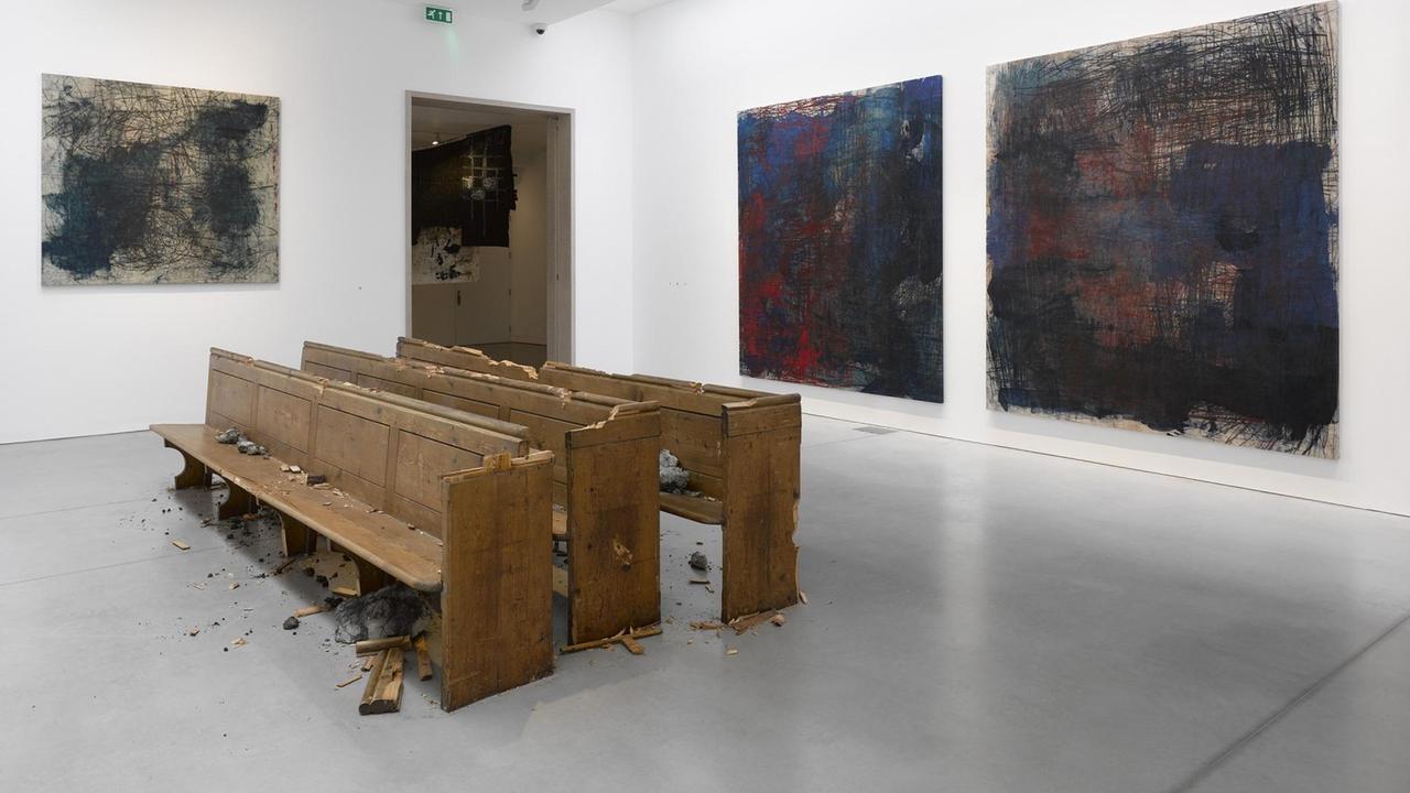 Kunstinstallation "Violent Amnesia" von Oscar Murillo. In einem Raum sind zerstörte hölzerne Sitzbänke und abstrakte Gemälde zu sehen.
