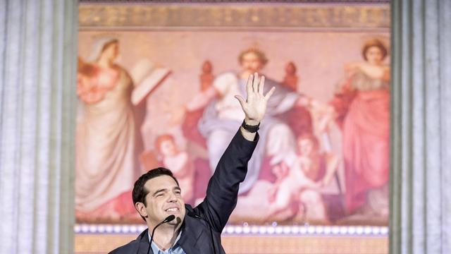 Alexis Tsipras reckt an einem Rednerpult zwischen zwei antiken Säulen vor einem historischen Gemälde die linke Hand in den Himmel.