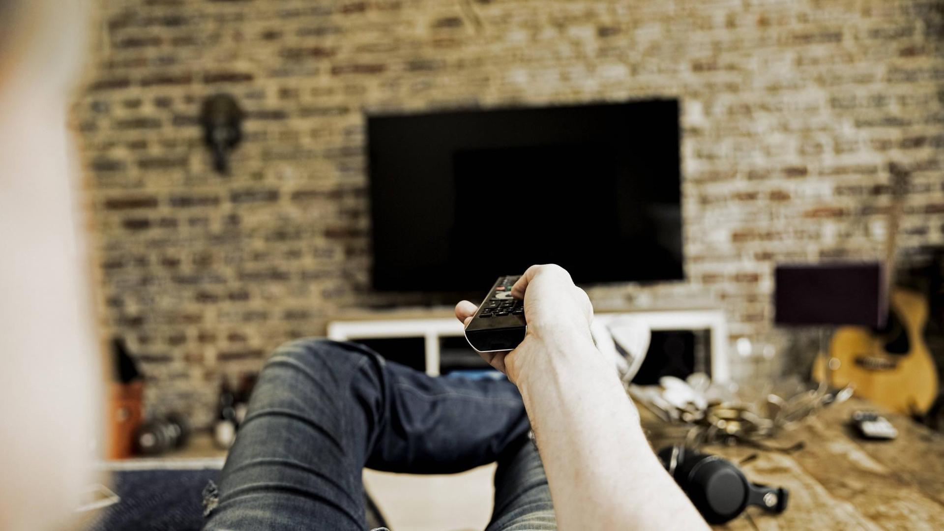 Ein Mann liegt auf einem Sofa und zeigt mit einer Fernbedienung auf einen Fernseher, der an der Wand angebracht ist Ein Mann liegt auf einem Sofa und zeigt mit einer Fernbedienung auf einen Fernseher, der an der Wand angebracht ist