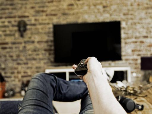 Ein Mann liegt auf einem Sofa und zeigt mit einer Fernbedienung auf einen Fernseher, der an der Wand angebracht ist