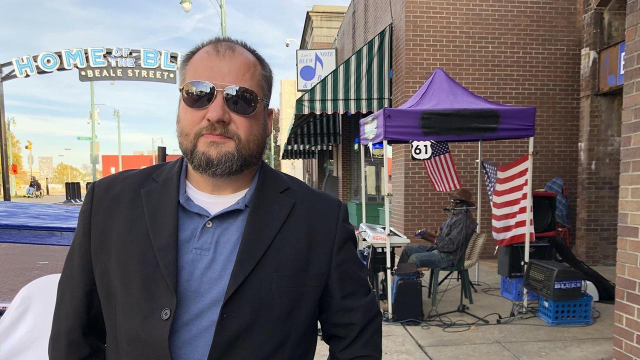 Ein Mann steht mit Sonnenbrille und im Sakko auf einer Straße. Ein Mann steht mit Sonnenbrille und im Sakko auf einer Straße.