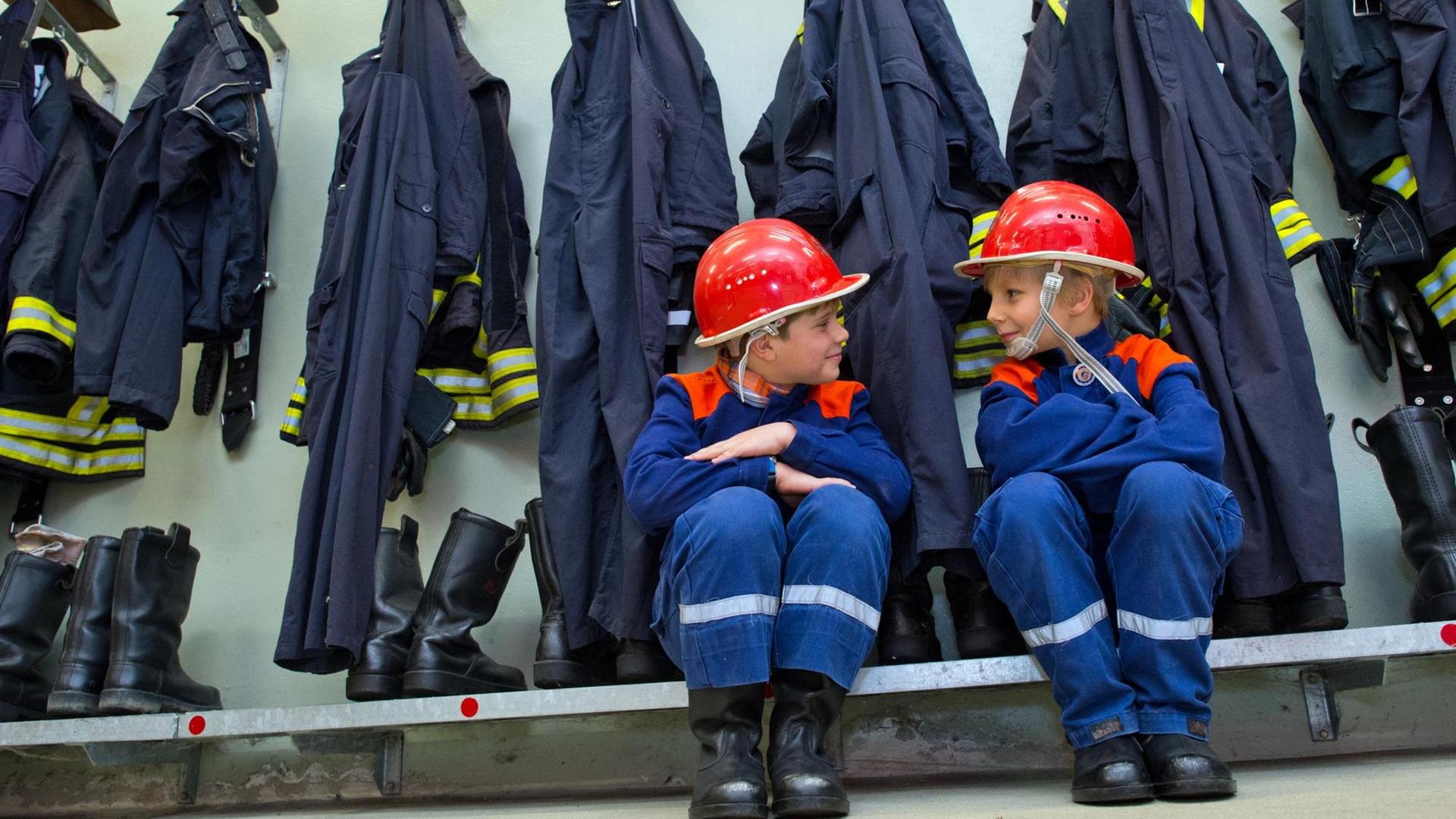 Zwei Jungen in Kinderuniform sitzen vor der ordentlich aufgehangenen Dienstkleidung der Freiwilligen Feuerwehr.