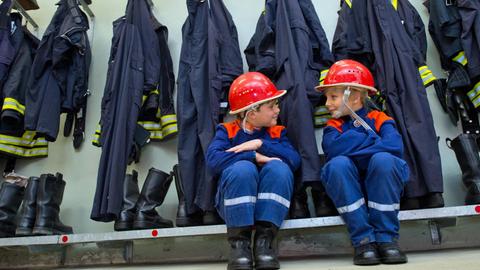 Zwei Jungen in Kinderuniform sitzen vor der ordentlich aufgehangenen Dienstkleidung der Freiwilligen Feuerwehr.