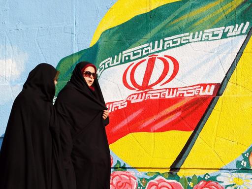Zwei iranische Frauen in Teheran vor einer iranischen Flagge, die als Graffiti an eine Wand gesprüht ist