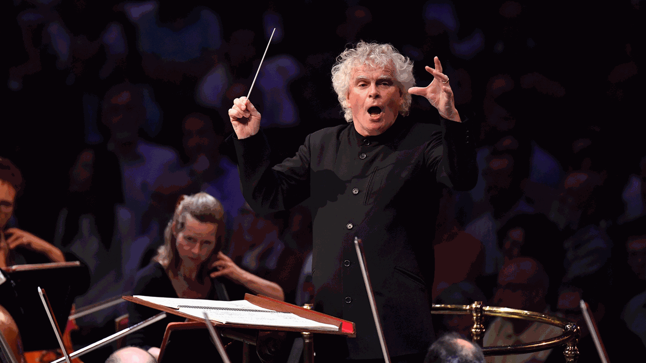 Ein Dirigent mit weißen Locken schwingt seinen Taktstock nach oben und animiert die Musiker mit aufreißendem Mund für einen Einsatz. Ein Dirigent mit weißen Locken schwingt seinen Taktstock nach oben und animiert die Musiker mit aufreißendem Mund für einen Einsatz.