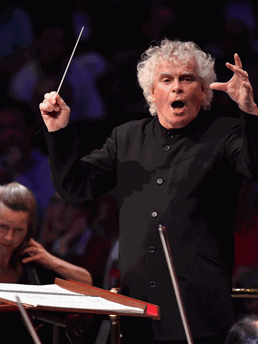 Ein Dirigent mit weißen Locken schwingt seinen Taktstock nach oben und animiert die Musiker mit aufreißendem Mund für einen Einsatz.