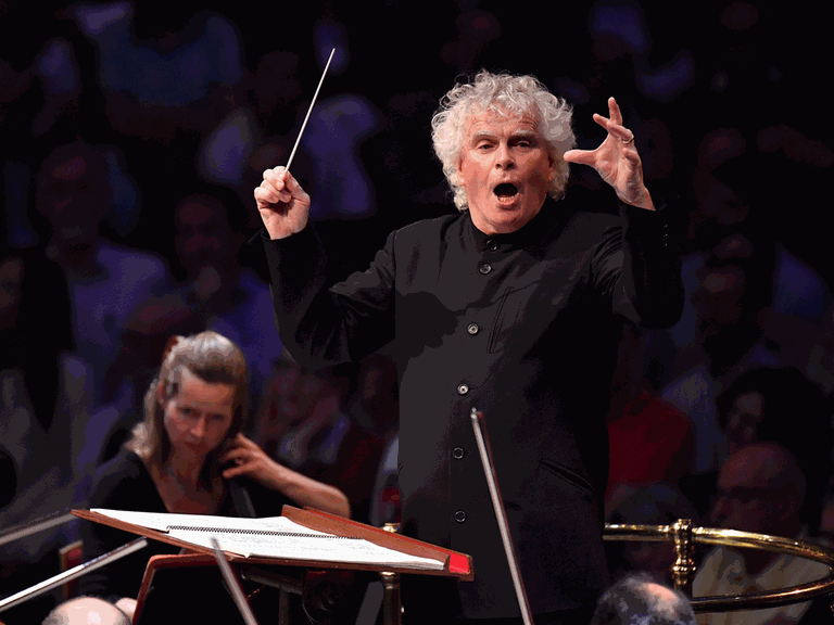 Ein Dirigent mit weißen Locken schwingt seinen Taktstock nach oben und animiert die Musiker mit aufreißendem Mund für einen Einsatz.