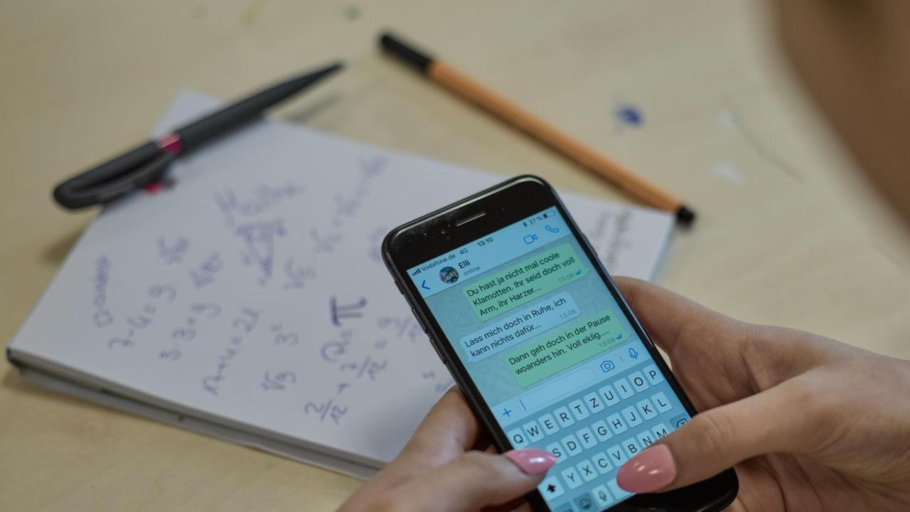 Auf einem Smartphone ist der Gesprächsverlauf einer Messenger-App zu sehen, dahinter ein Schulheft und Stifte.