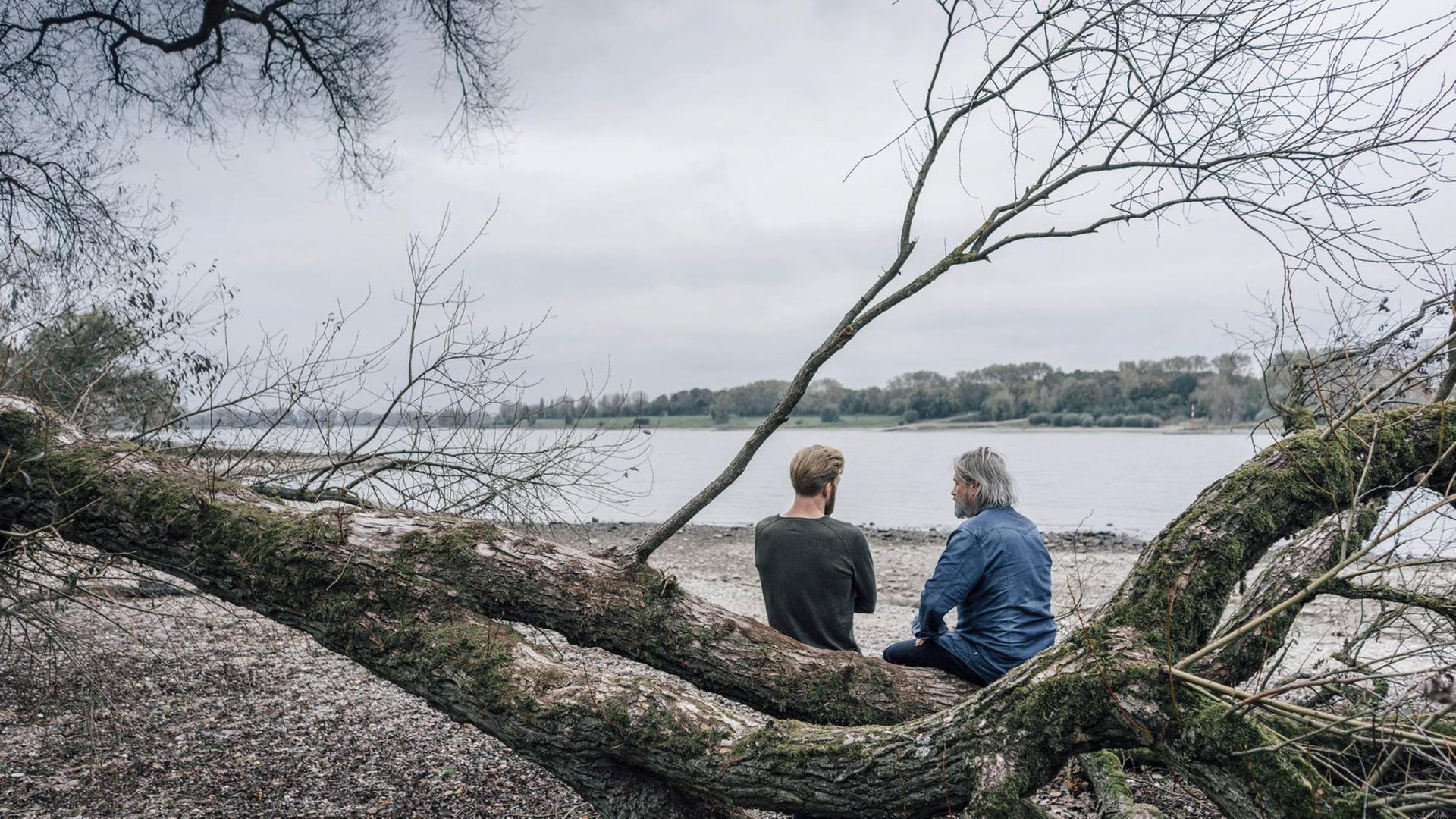 Vater und Sohn sitzen auf einem umgefallenen Baum an einem Fluss und unterhalten sich.