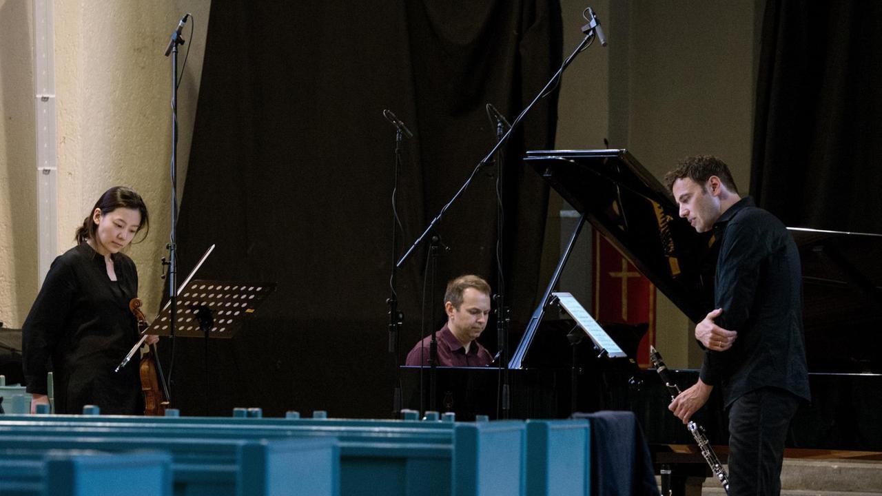 Eine Geigerin, ein Pianist und ein Klarinettist stehen in einem Kirchenraum von Mikrophonen umgeben und schauen konzentriert auf ihre Noten. Eine Geigerin, ein Pianist und ein Klarinettist stehen in einem Kirchenraum von Mikrophonen umgeben und schauen konzentriert auf ihre Noten.
