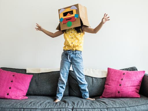 Ein Kind springt auf einem Sofa. Über dem Kopf trägt es einen Pappkarton mit einem bunten Gesicht.